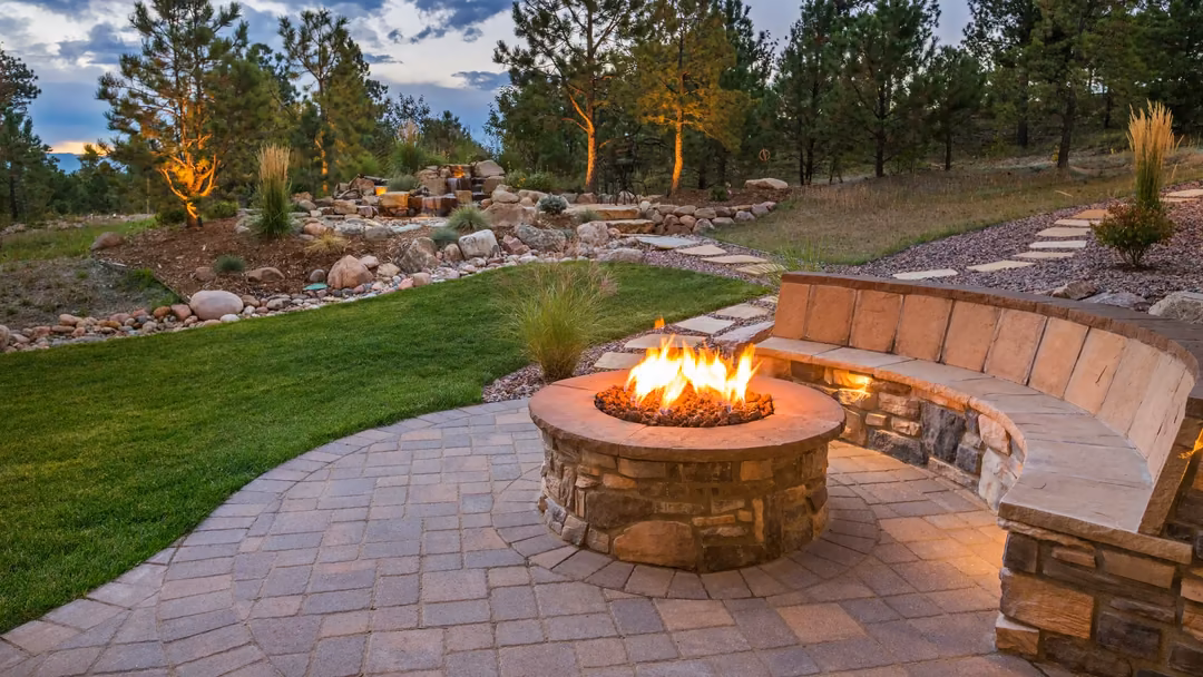 Stone fire pit with curved seating wall on a paver patio, surrounded by landscaped beds and accent lighting at dusk