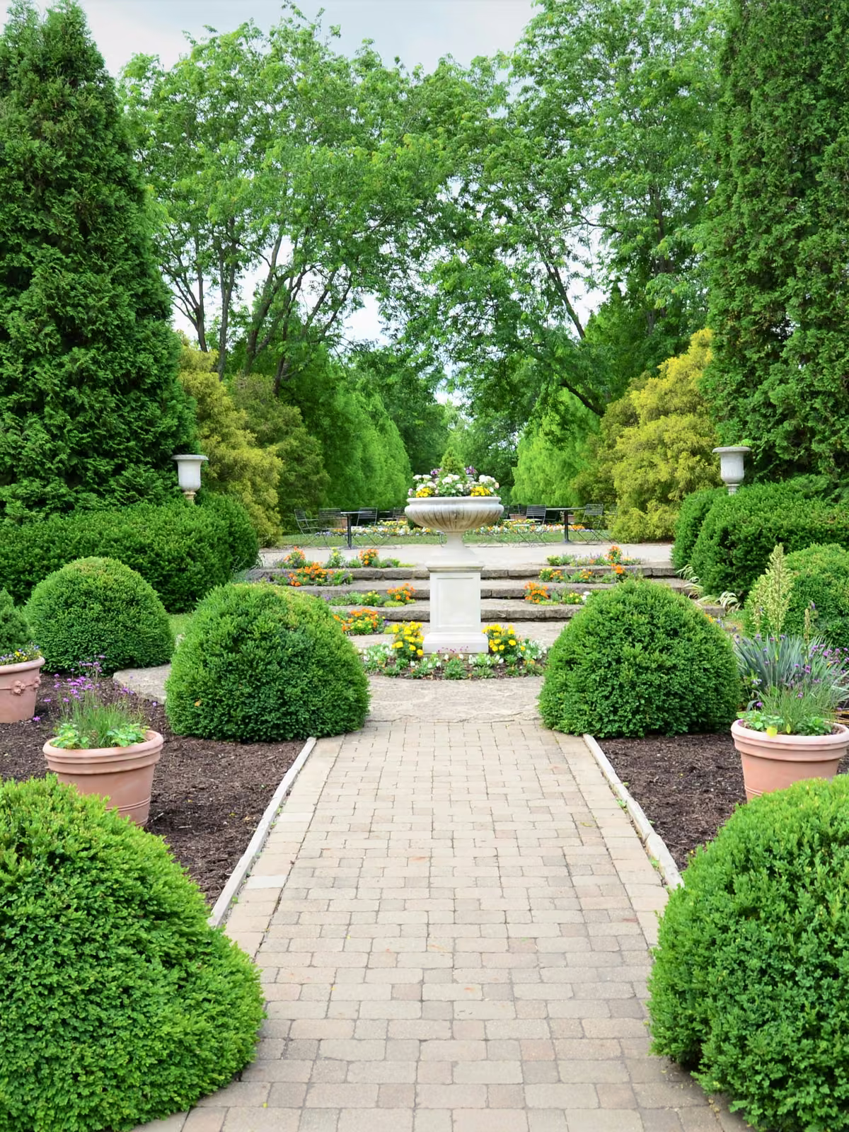 Formal garden with a paver walkway, clipped boxwood spheres, and stone urn planters